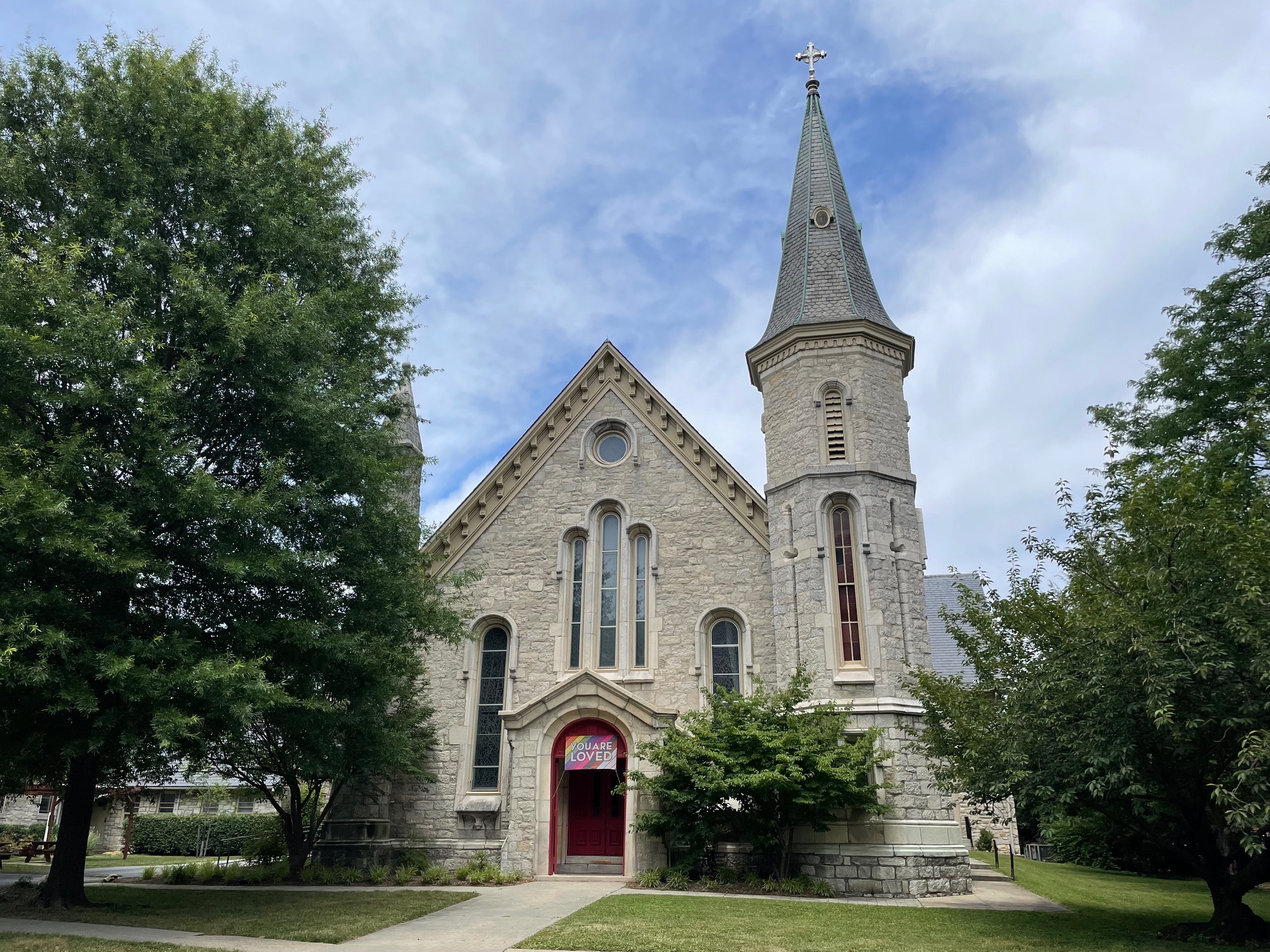 A historic stone church with a tall steeple stands surrounded by lush trees, under a partly cloudy sky, featuring a welcoming red door.