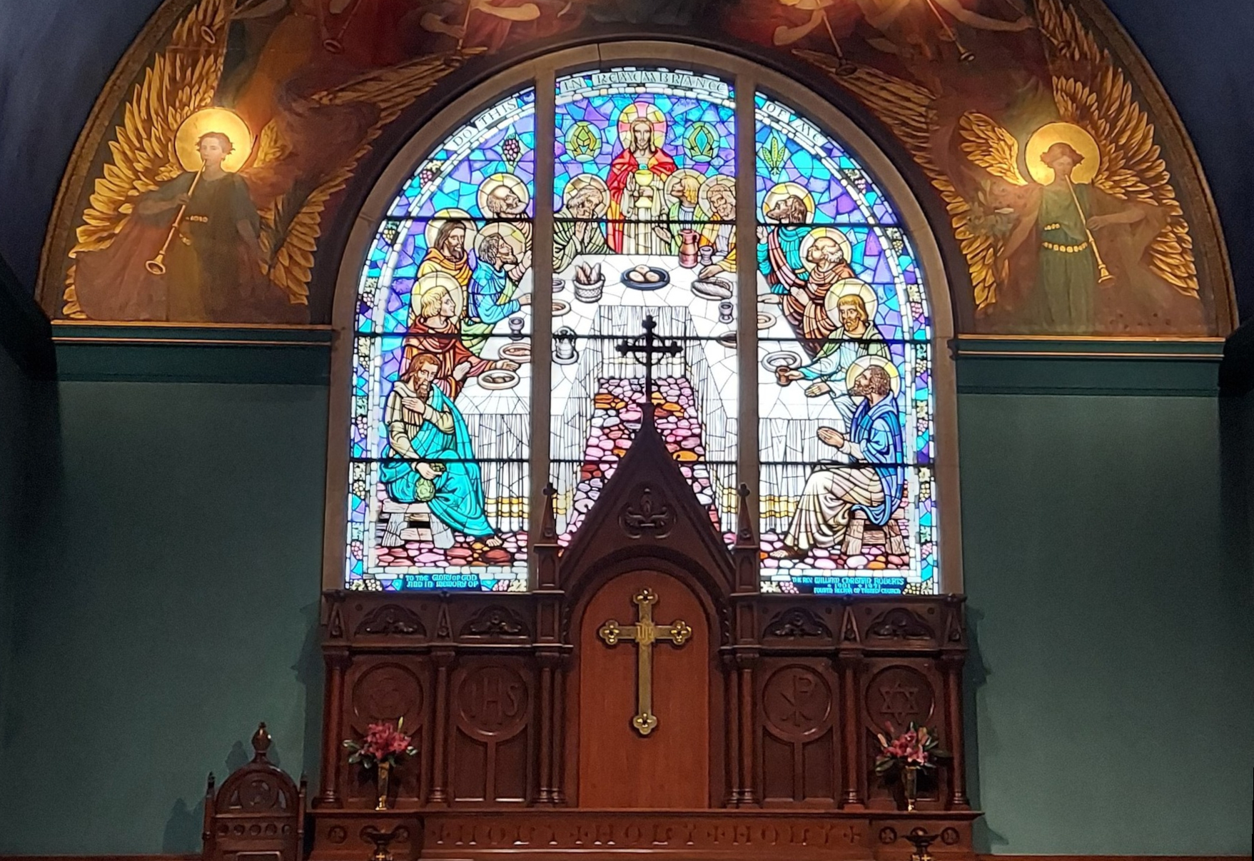 Stained glass window depicting the Last Supper above a wooden altar with a cross, flanked by angel figures in a church interior.