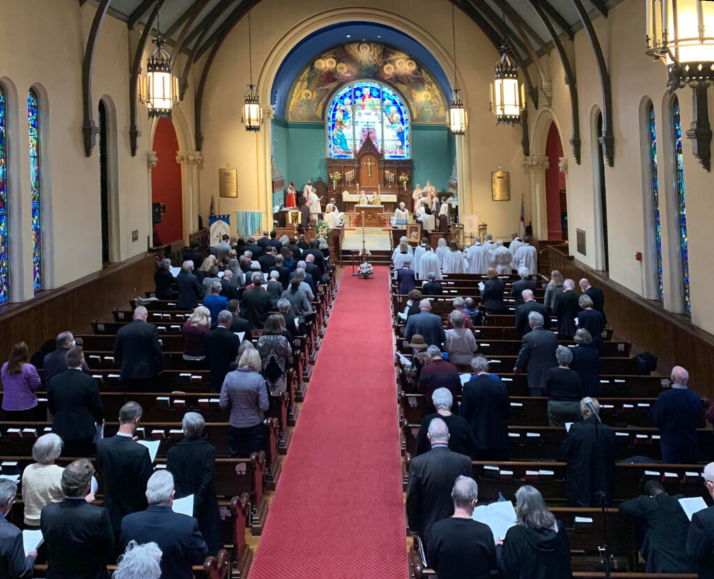 Inside a church, people are attending a service. The interior features stained glass windows and a red carpet aisle leading to the altar.