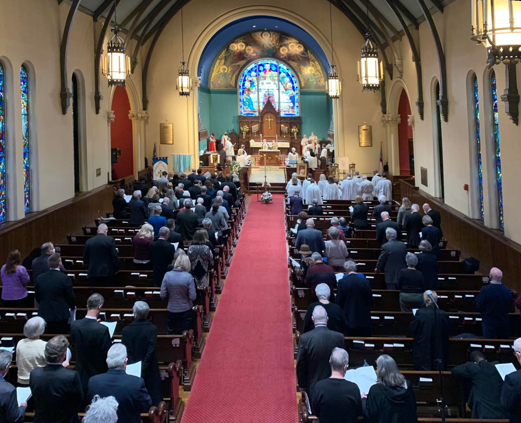Inside a church, people are attending a service. The interior features stained glass windows and a red carpet aisle leading to the altar.