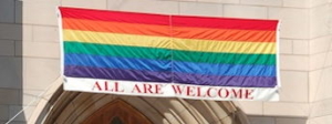 A rainbow flag with "All Are Welcome" hangs outside a stone building, possibly a church, emphasizing inclusivity and acceptance.