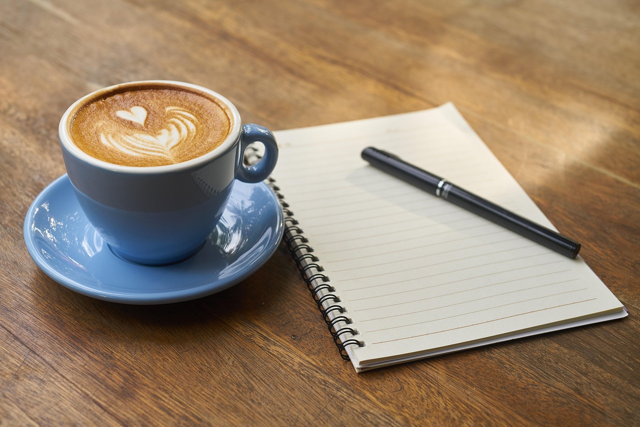 A blue cup of latte art sits on a wooden table beside an open lined notebook with a pen.
