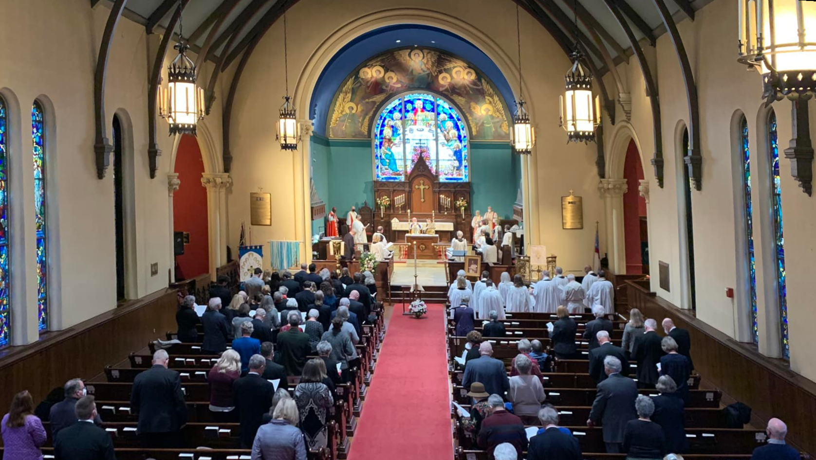 Congregation gathers inside a church with stained glass, arched ceiling, red carpet, and clergy at the altar. People are seated in pews.