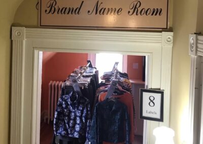 A room with designer clothes on racks, labeled "Brand Name Room." Warm lighting, vintage decor, and a sign reading "8 Labels" are visible.