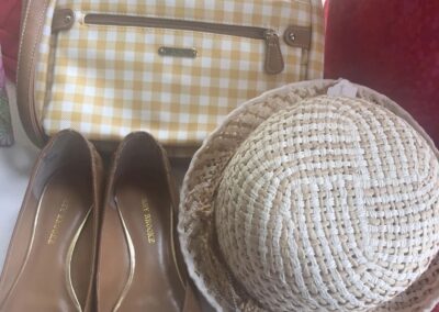A yellow checkered handbag, straw hat, and brown flats with gold emblem displayed on a table with red and pink fabrics nearby.