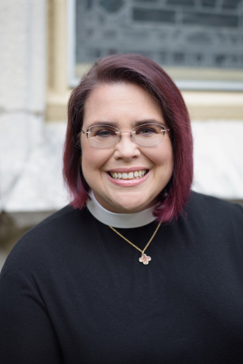 A smiling person with glasses wearing a clerical collar, standing in front of a light-colored, textured wall, outdoors.