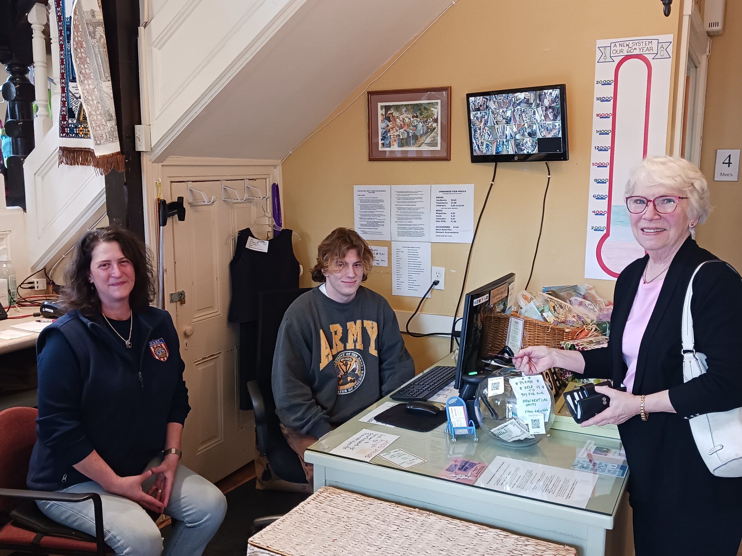 Three people in a cozy room near a counter. A TV displays security footage, and a thermometer chart is on the wall.