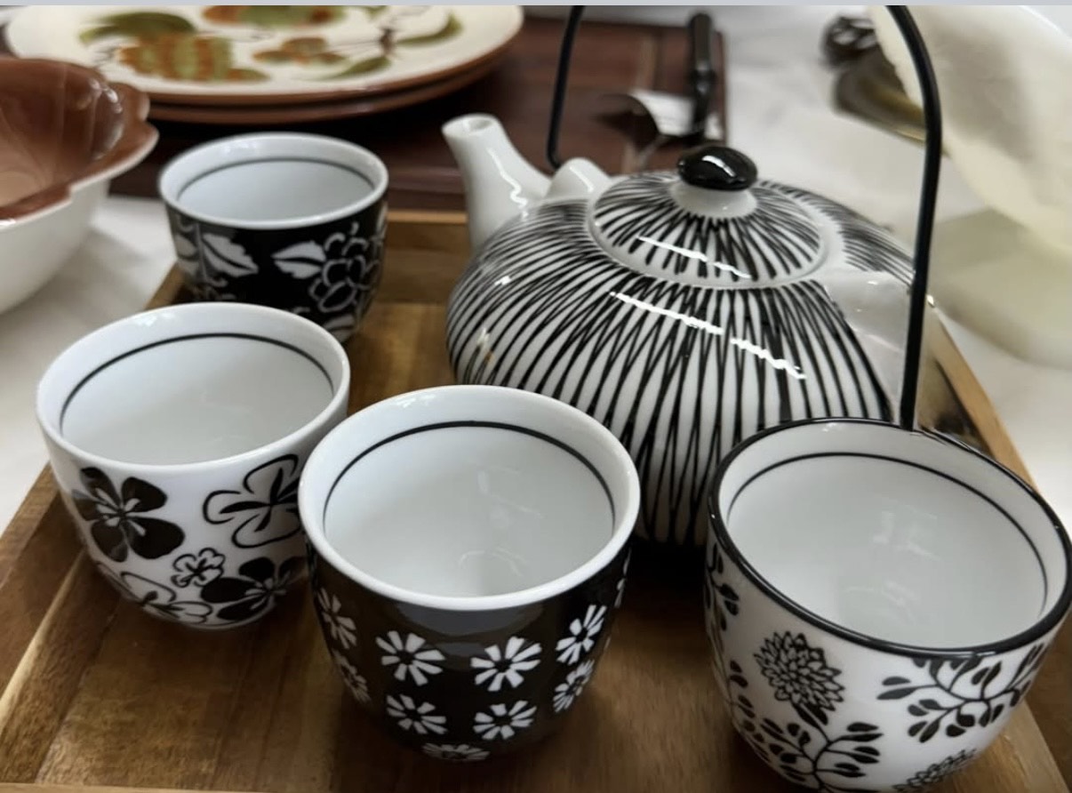A wooden tray holds a black-and-white teapot with four matching floral teacups, surrounded by various ceramic dishes on a table.