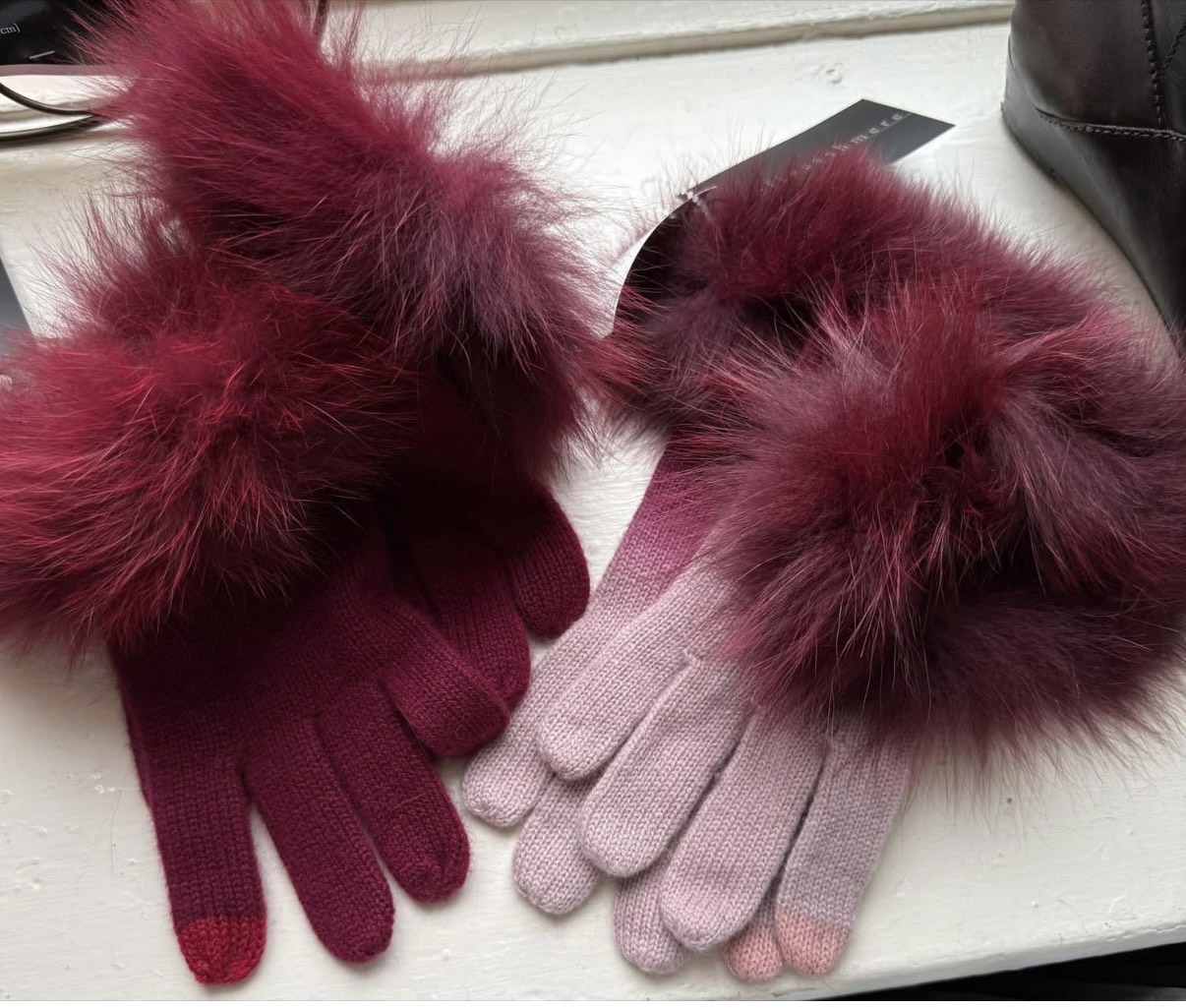 Two pairs of gloves with furry cuffs, one burgundy and one pink, displayed on a white surface beside a dark-colored object.