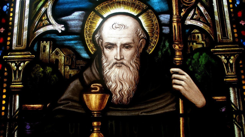 Stained glass depicting a bearded person with a halo, holding a staff and cup, set against an architectural backdrop with medieval buildings.