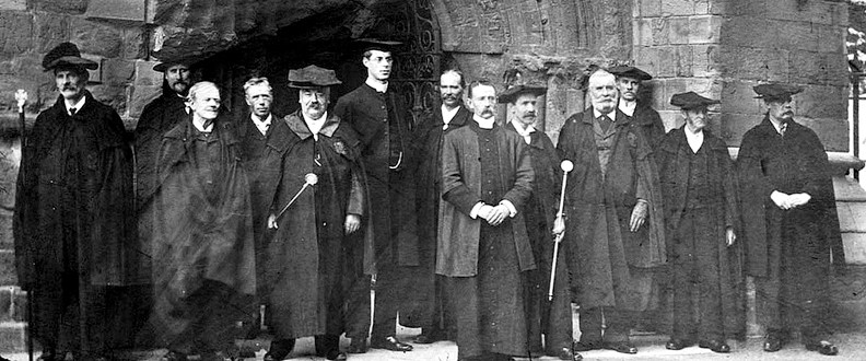 A group of people in academic robes stands outside a historical stone building, likely a university, holding staffs, posing for a photograph.