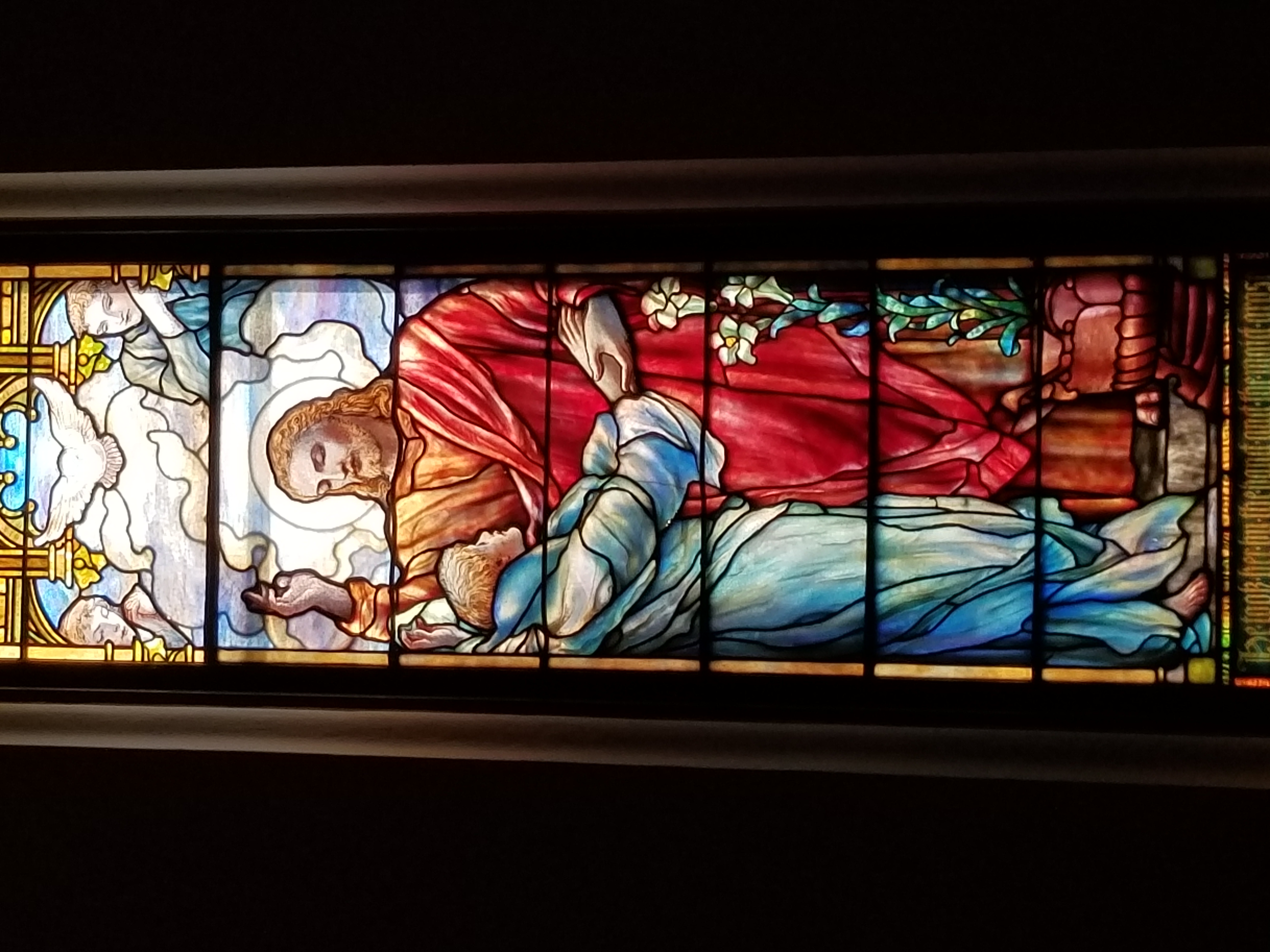 Stained glass depicting religious figures, including a haloed person in a red robe, angelic figures, lilies, and a dove at the top.