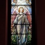 A stained-glass window depicts an angelic figure with wings and colorful robes, surrounded by cherubs and intricate details. Rich colors and ornate design.
