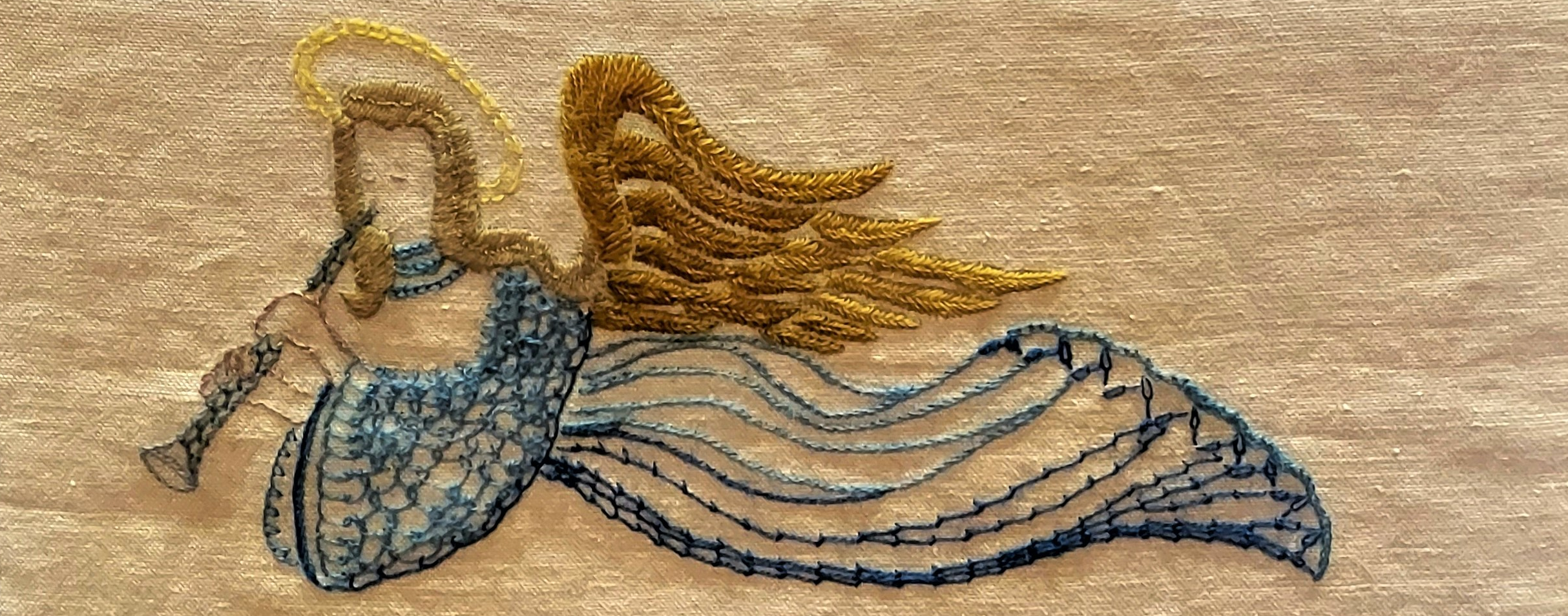 Embroidered angel with golden wings and halo, playing a trumpet, on fabric background, using blue and gold threads for intricate detailing.