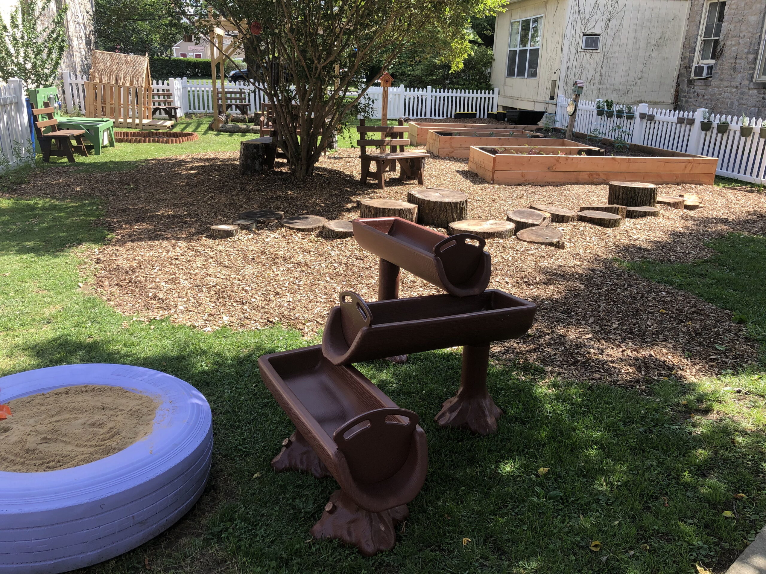 Outdoor play area with wooden structures, a sandpit, and log steps. Surrounded by a white picket fence and green grass.