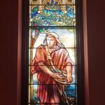 Stained glass window depicting a person in ancient attire sowing seeds. Above is a decorative architectural design with trees and blue sky surrounding.