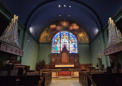 A church interior with wooden pews, pipe organ, stained glass window, and ornate altar featuring a cross and religious artwork.