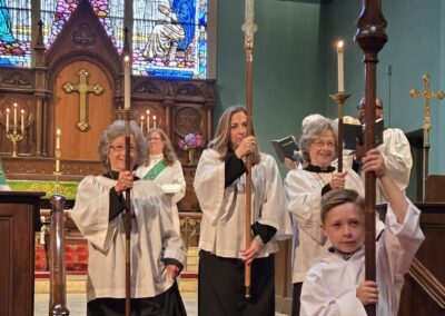 Religious procession inside a church, featuring stained glass windows, wooden cross, and people wearing white robes, amid candlelight and sacred atmosphere.