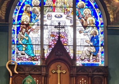 A person in green robes stands before an altar, with a stained glass window depicting the Last Supper in the background.