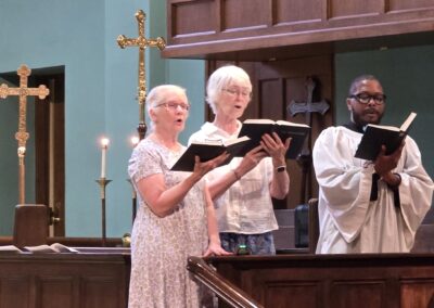 Three people singing in a church setting, holding books, flanked by ornate crosses, with candles in the background.