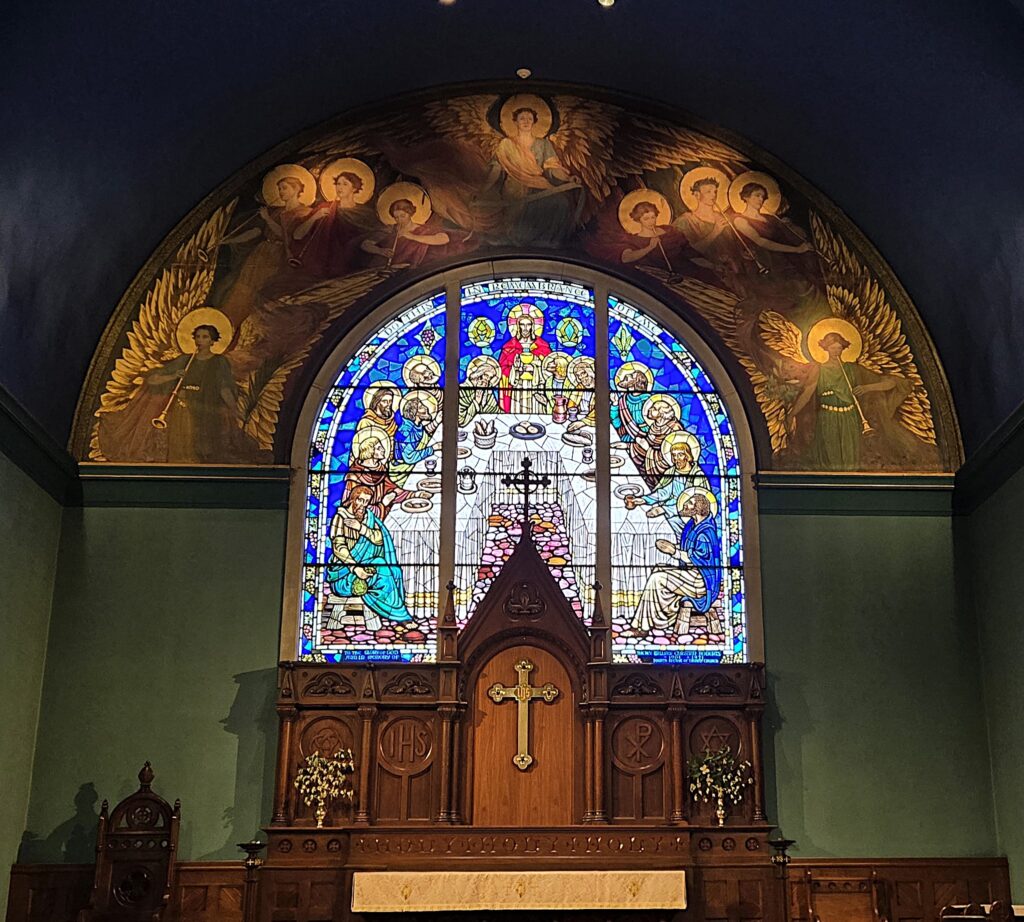 Stained glass window depicting religious figures above a wooden altar with a cross, framed by angel paintings in a chapel setting.