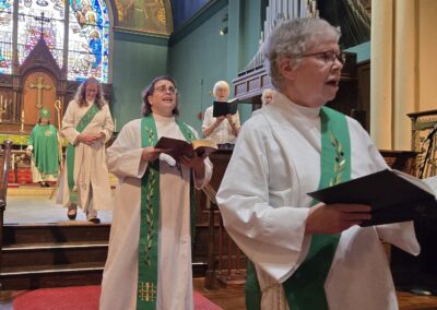 Religious service with people in robes, holding books and singing in a church. Stained glass window and organ pipes are visible.