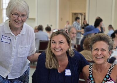 Three people smiling at a gathering with others in the background, wearing name tags in a warmly lit indoor setting.