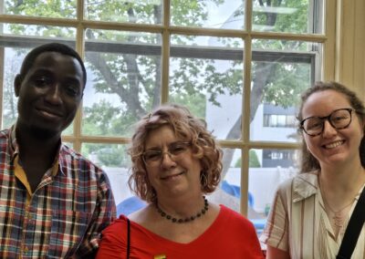 Three people smiling indoors in front of a window with trees and buildings visible outside, creating a casual and friendly atmosphere.