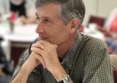 A person in a plaid shirt sits with hands clasped, looking thoughtful. Blurred people and flowers are visible in the background.