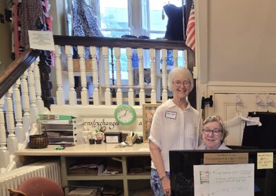 Two people smiling at a consignment store counter, surrounded by clothes and decor. Small American flag displayed.
