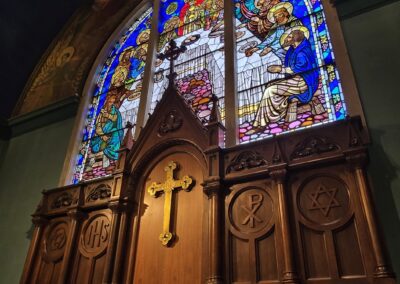 Stained glass window in a church depicts religious figures, with intricate woodwork featuring a golden cross and symbolic carvings below.