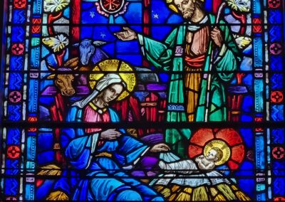 Stained glass artwork depicting nativity scene, with two adults and baby surrounded by farm animals and symbolic motifs on a vibrant blue backdrop.