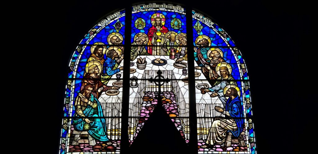 Stained glass window depicting the Last Supper with vibrant colors, featuring religious figures around a table, inside a church setting.