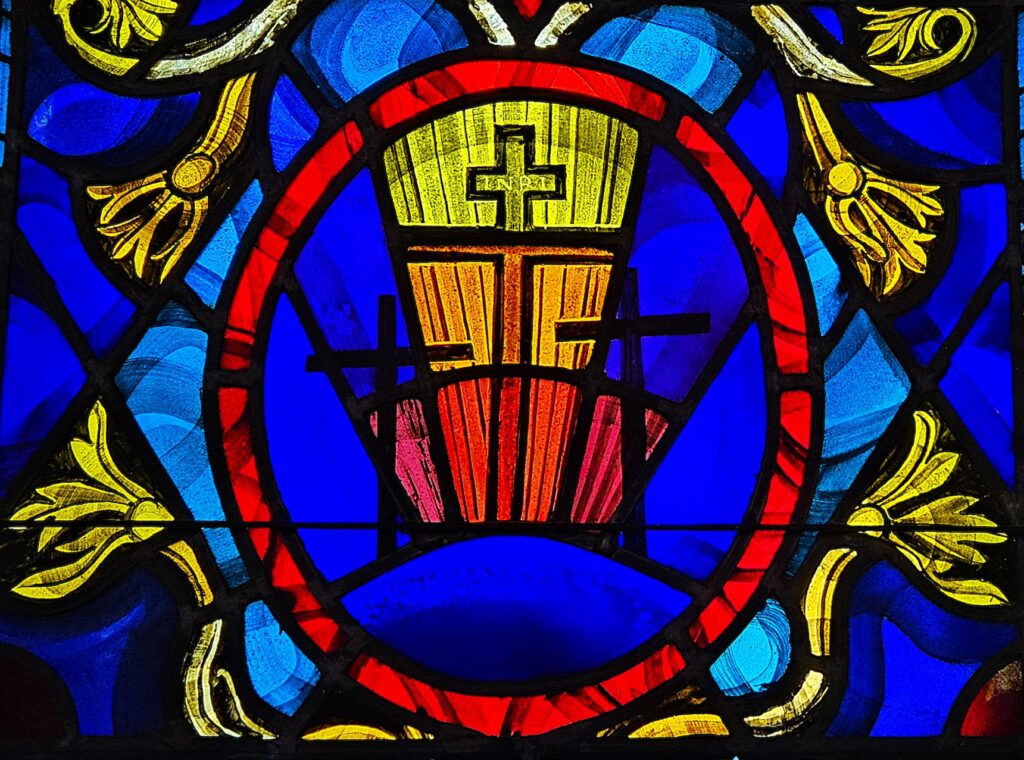 Stained glass window with vibrant blue, red, and yellow colors, featuring a cross design and floral patterns, symbolizing religious themes.