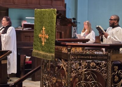 Three people in a church setting, reading from books, with ornate wooden furnishings and embroidered green cloth.