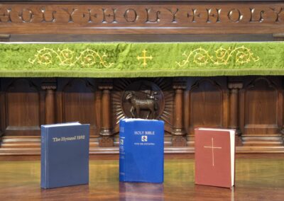 Ornate wooden altar with "Holy" engraving, three religious books displayed: The Hymnal 1982, Holy Bible, red book, featuring a green altar cloth.