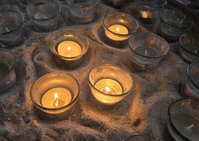 Small glass candle holders with lit and unlit candles are arranged on sand, creating a serene and contemplative atmosphere.