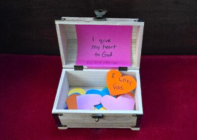 A small wooden box contains colorful paper hearts with religious messages, placed on a red surface, suggesting themes of faith and devotion.