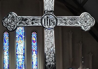 An ornate cross is displayed in front of colorful stained glass windows inside a church, featuring intricate floral and symbolic designs.