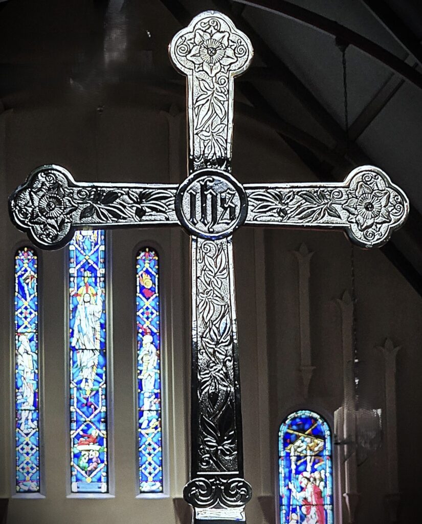 An ornate cross is displayed in front of colorful stained glass windows inside a church, featuring intricate floral and symbolic designs.