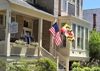 Charming stone house with U.S. and Maryland flags, flower boxes, and clothing display. "The Surprise Shop" sign indicates Trinity Episcopal Church affiliation.