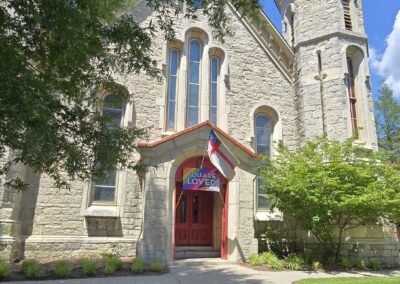 Stone church with arched windows, red doors, and a "You Are Loved" sign. Flags displayed. Trees partially shade the building entrance.