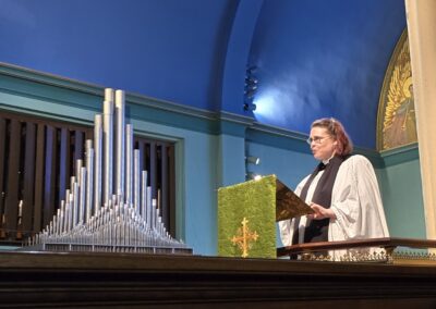 A person in religious attire stands at a lectern beside large organ pipes within a brightly lit church interior.