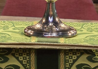 A silver chalice on an altar with green and gold fabric in a church with rows of wooden pews and red carpet.