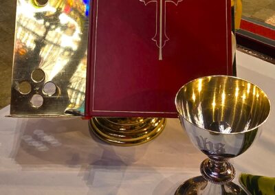 A red book with a cross, silver chalice, and golden reflection are arranged on a table inside a church setting.
