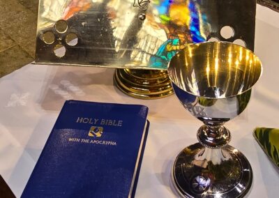 Chalice, Bible, and decorated metal piece on altar, featuring colorful reflections from stained glass in a religious setting.