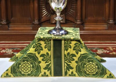 A silver chalice sits on an elaborately decorated altar with green and gold embroidered cloth, beneath a wooden structure inscribed with "Holy."