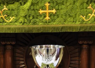 An ornate silver chalice rests on an altar with green and gold embroidered cloth, beneath a carved wooden panel labeled "HOLY."