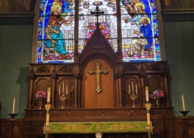 An ornate church interior with a stained-glass window, wooden altar featuring a cross, candles, and decorative cloth, emanating a serene, historic ambiance.