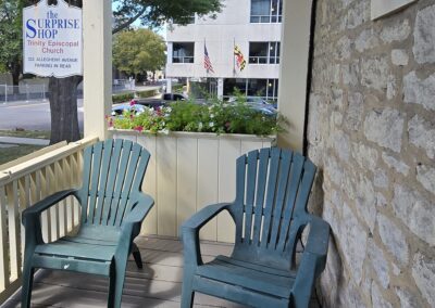 A porch with two chairs and a sign for The Surprise Shop, Trinity Episcopal Church. Two flags are visible across the street.